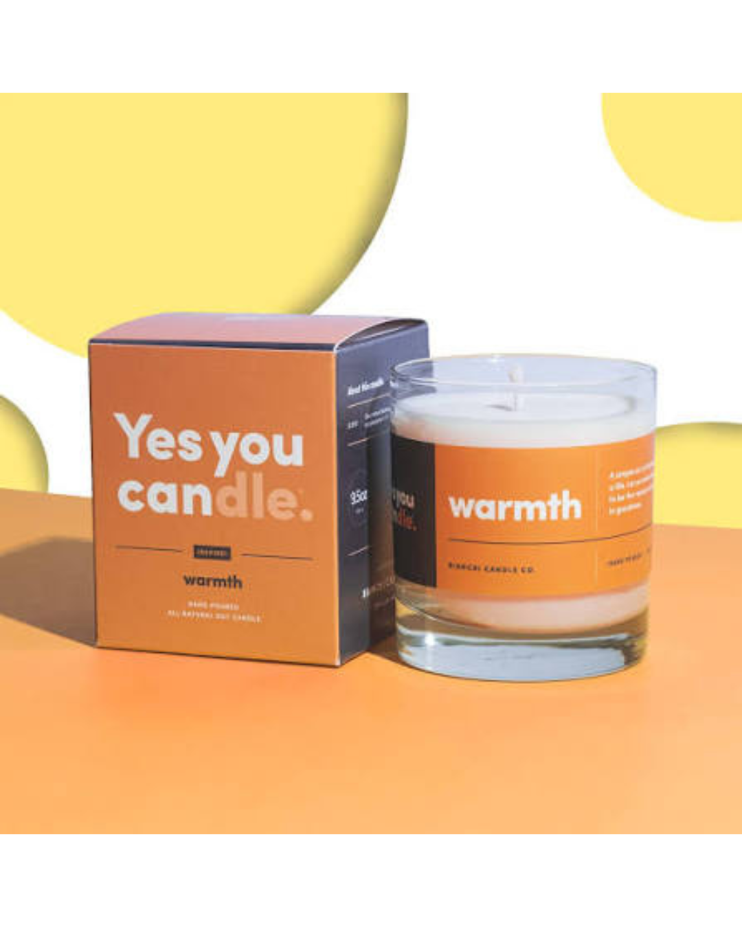 A glass jar candle with white wax and an orange label next to its matching orange and dark gray box, branded 'warmth' and 'Yes you candle.' on a bright orange and yellow background.
