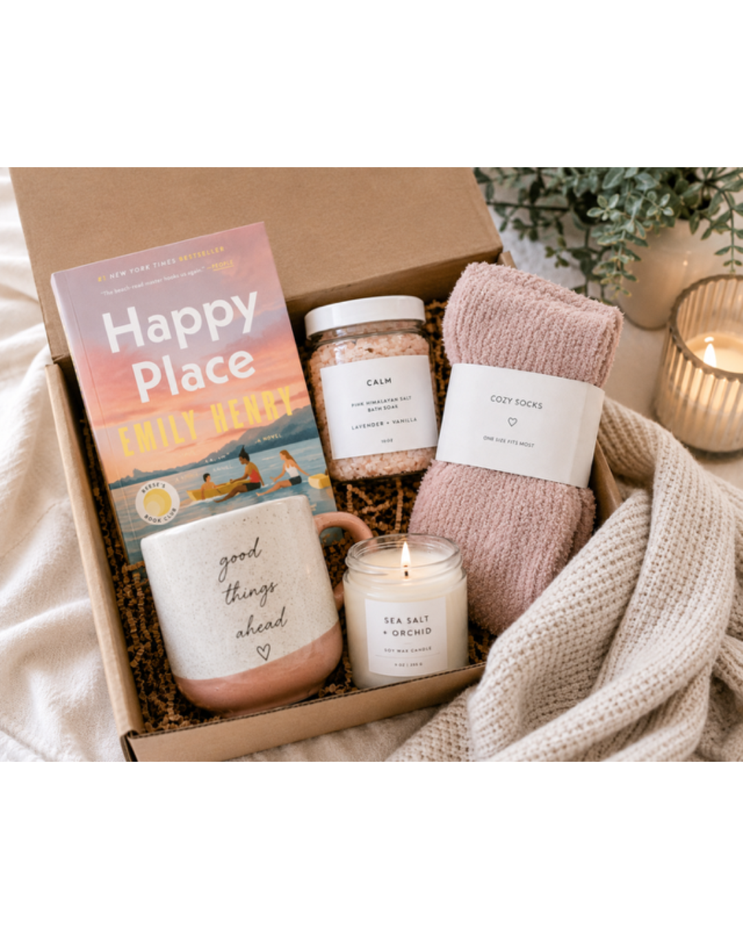 A curated gift box set containing a 'Happy Place' journal, a 'good things about' mug, a jar of bath salts, a candle, and cozy pink socks, arranged with a cream knit blanket and a lit candle in a styled setting.
