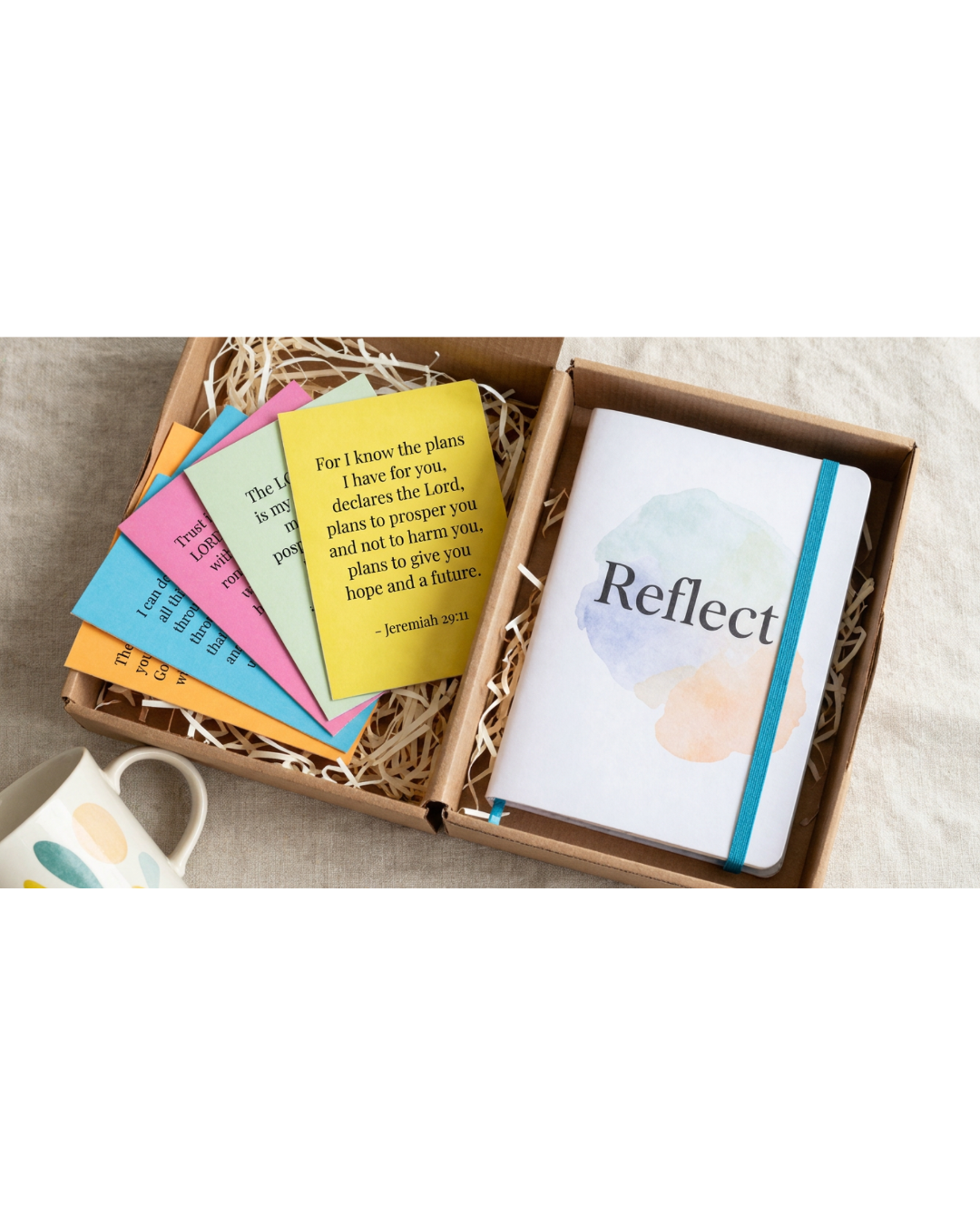 A gift set in a brown cardboard box containing a white journal with a blue elastic band and the word 'Reflect' printed on the cover, alongside several colorful sticky notes in pink, blue, yellow, and orange.
