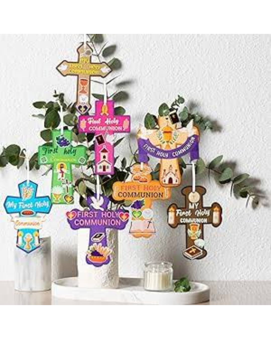A decorative display of colorful wooden First Holy Communion crosses and signs arranged in a white vase with greenery, set on a white tray against a light textured wall.