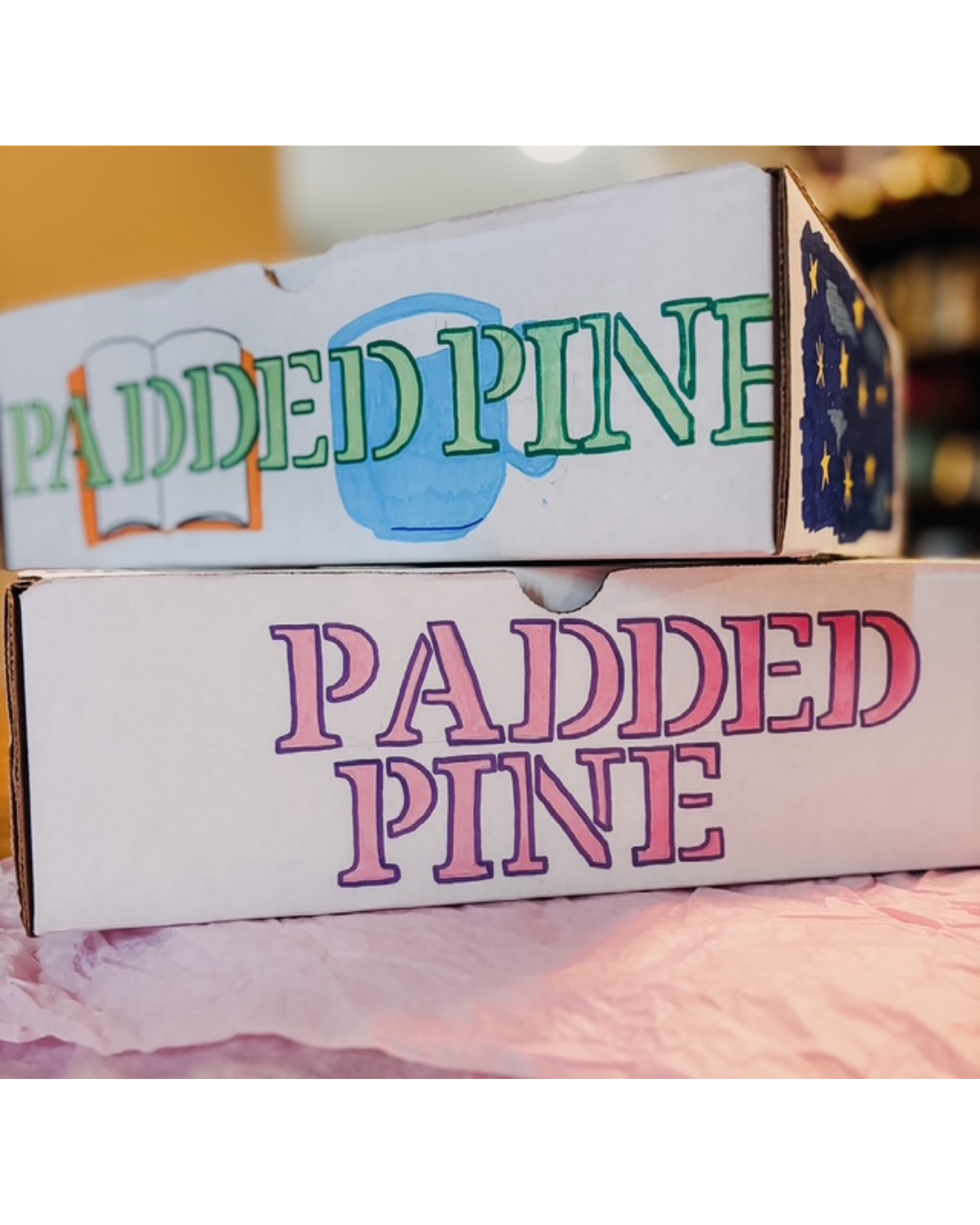Two white cardboard boxes stacked on pink tissue paper, both printed with the words 'PADDED PINE' in large outlined letters. The top box has green letters and a blue book illustration, while the bottom box has pink letters with a purple shadow effect.