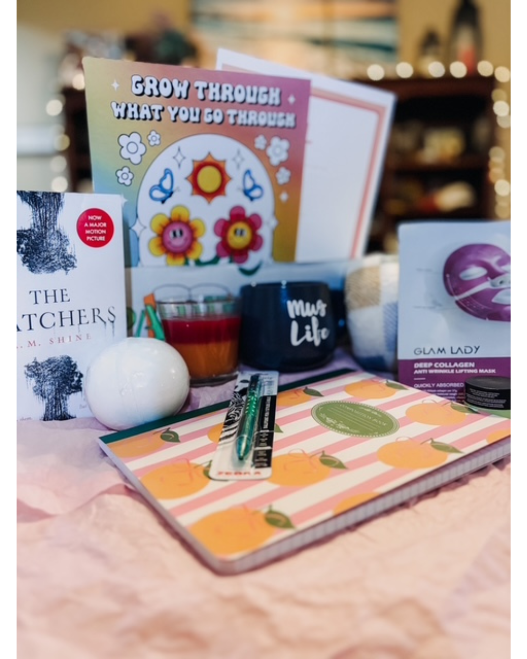 A styled gift basket or curated product arrangement on a pink fabric surface, featuring a notebook with orange and pink stripes, a black mug with white text, a book, a card with colorful flowers, a candle, and a boxed sheet mask. The background is softly blurred with warm lights.