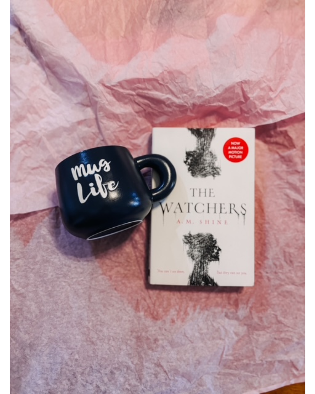 A black ceramic mug with 'my life' written in white script, placed next to a copy of the book 'The Watchers' by A.M.