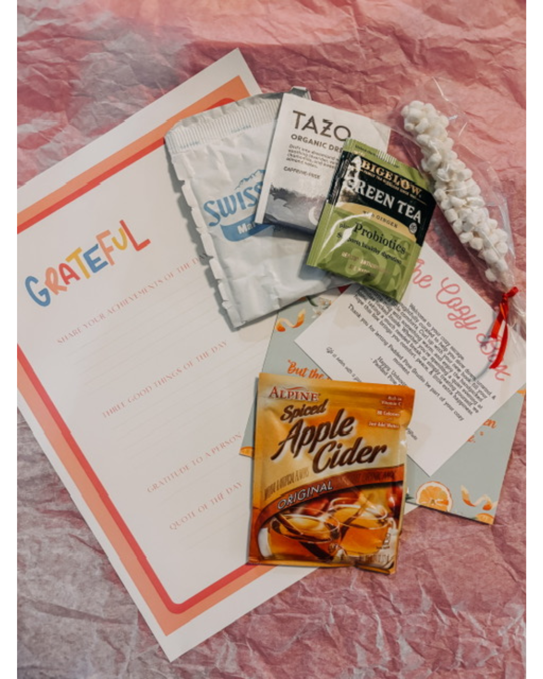 A flat-lay arrangement of gift items on a crinkled pink background, including a printed gratitude card, Sweetener Swap cards, Tazo tea bags, a green herbal tea packet, an Altini Apple Cider packet, and a white beaded bracelet.