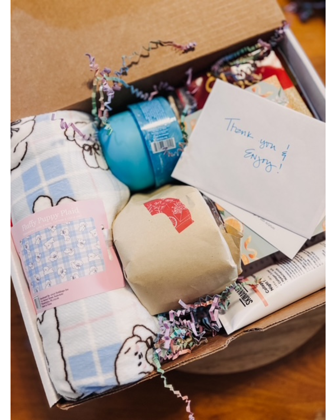 An open cardboard gift box filled with assorted items, including a blue round container, a beige wrapped pouch with a red logo, a folded white and blue patterned textile, a pink card, and a handwritten white note that says 'Thank you so much!'.