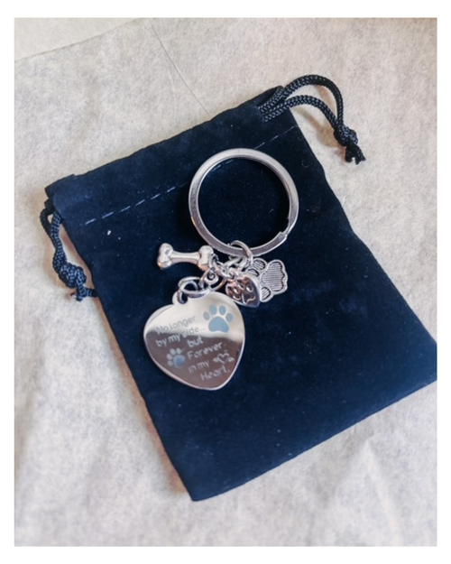 A silver metal keychain with engraved heart-shaped charm, paw print, and bone charms, displayed on a black velvet drawstring pouch over a light beige fabric background.