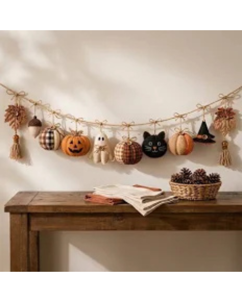 An autumn-themed fabric garland hanging on a wall above a rustic wooden table. The garland features various seasonal decorations including pumpkins, acorns, a ghost, a black cat, a witch hat, and tassels, all attached to a natural jute string.