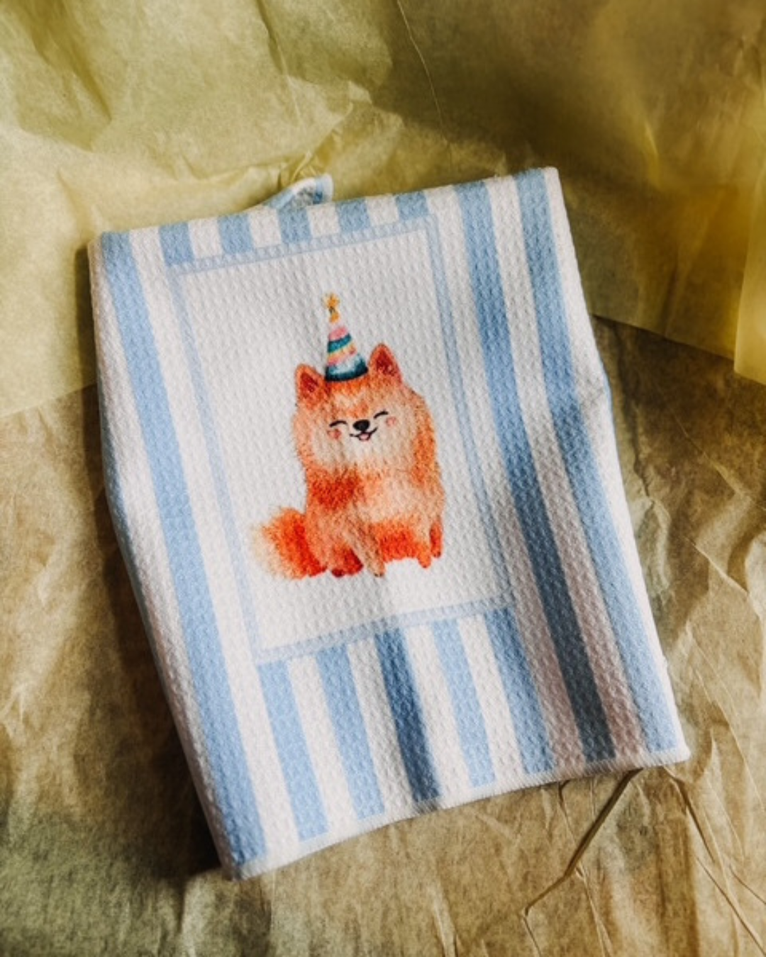 A folded waffle-weave kitchen towel with light blue and white stripes, featuring a watercolor illustration of an orange fluffy dog wearing a party hat, displayed on a textured brown paper background.