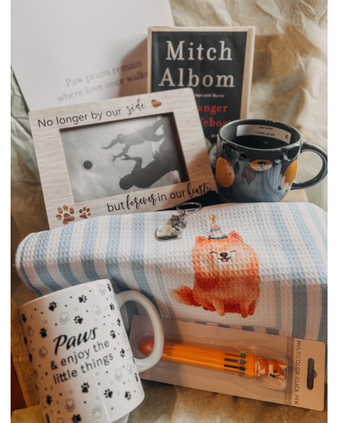 A curated gift bundle featuring a blue and white striped waffle-knit blanket with a cartoon dog illustration, a white mug with black paw prints and the text 'Paws & enjoy the little things', a wooden picture frame, a black mug with cat faces, a book titled 'Mitch Albom', and a small orange item in a blister pack.