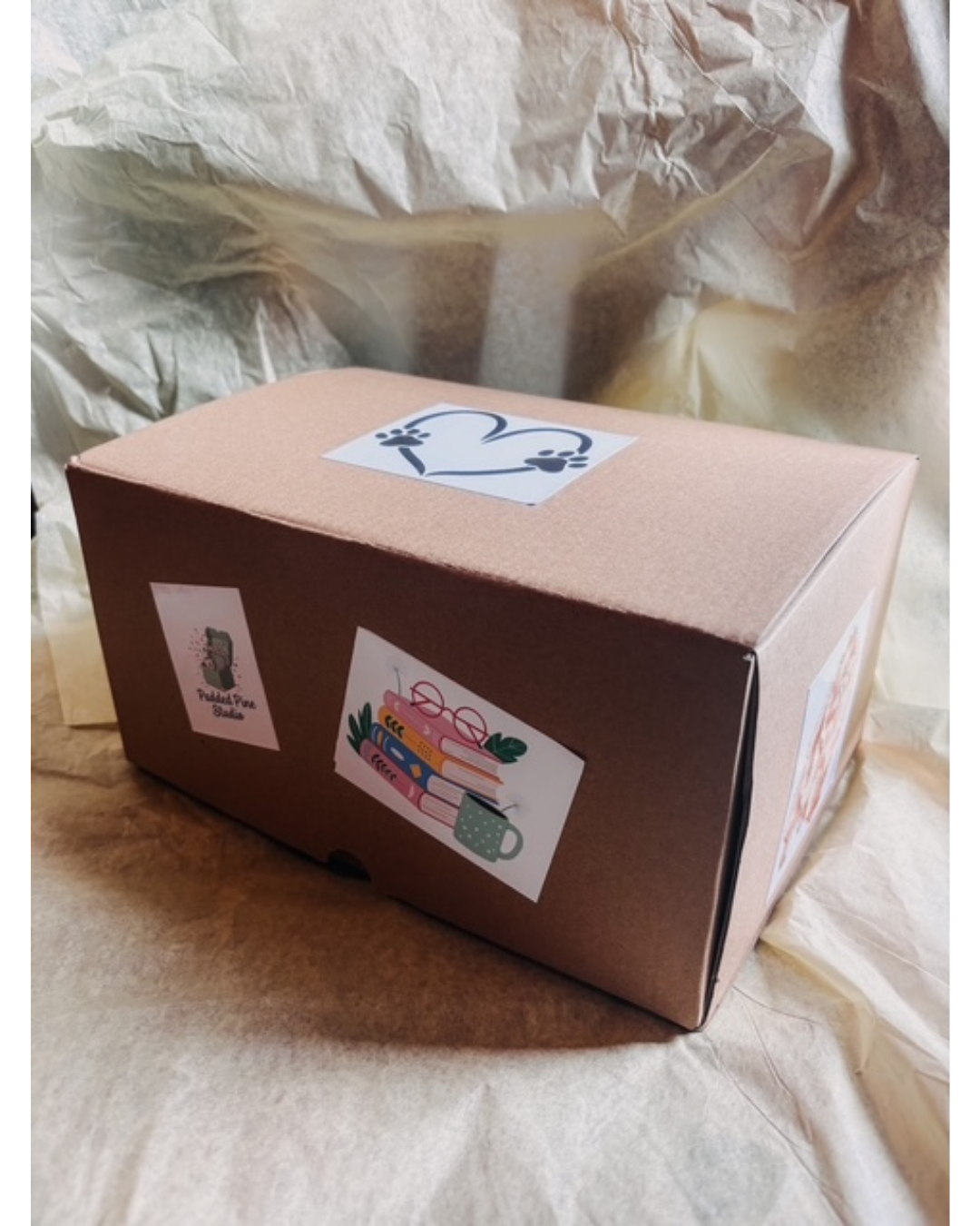 A brown cardboard shipping box with several decorative stickers on the top and sides, placed on beige tissue paper.